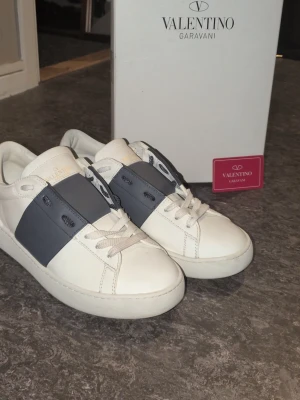 Valentino Garavani vita sneakers - Snygga vita sneakers från Valentino Garavani med bred mörkgrå rem över mitten. Klassisk låg modell med vita skosnören och diskret logga på tungan. Tillverkade i läder för en lyxig känsla och stilren look.