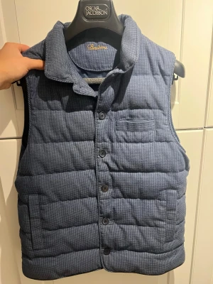 Blå dunväst från Stenströms  - Snygg blå dunväst från Stenströms med diskret rutmönster. Västen har knäppar framtill, två sidofickor och en liten bröstficka. Perfekt för lager-på-lager och ger en stilren look. Fylld med dun för extra värme och komfort. Köpt för 3500