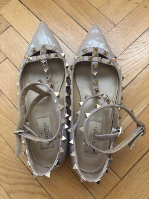Valentino Rockstud ballerinaskor beige - Ballerina skor från Valentino i beige lack med Rockstud-nitar. Snygga inför sommaren. Köpte från Vestiaire Collective och har äkthetsbevis. Om du är osäker på äktheten kan du köpa från min Vestiaire då man kan lägga till äkthetsbevis, den har däremot redan gjort det. Kostar ca 8000kr nypris men jag säljer mycket billigare. Små defekter: litet svart märke på vänster sko (se bild), lite läder är borta på topparna av skorna, lite rynkor på höger sko (se bild). Kommer med röd valentino skolåda.