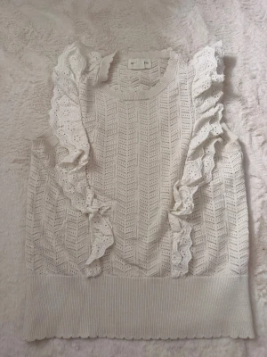 Vit ärmlös topp med volangdetaljer - Supersöt vit ärmlös topp med broderade volangdetaljer längs axlarna och ett fint hålmönster över hela plagget. Ribbad kant nedtill och runt halsen ger en extra touch. Perfekt för dig som gillar romantisk och feminin stil.