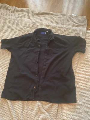 Svart kortärmad skjorta Jack & Jones - Svart kortärmad skjorta från Jack & Jones med klassisk krage och knäppning framtill. Skjortan är tillverkad i ett lätt och skönt material, perfekt för varma dagar. Enkel och stilren design som passar till många olika outfits.