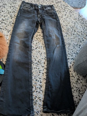 Svarta bootcut jeans med kontrastsömmar stl 164 - Säljer ett par svarta bootcut jeans med slitningar och vita kontrastsömmar. Klassisk femficksmodell och knappgylf. Jeansen har en snygg tvättad look och passar perfekt till en avslappnad stil.