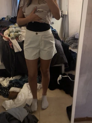Vita höga shorts med knapp - Snygga vita shorts med hög midja och en brun knapp framtill. Klassisk och enkel design som passar till många olika stilar. Perfekta för varma dagar och lätta att matcha med olika toppar.