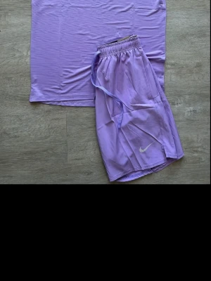 Lila Nike shorts med snörning - Säljer ett par lila shorts från Nike med resårmidja och snörning i midjan. Shortsen har en sportig look och är perfekta för träning eller chill. Nike-logga i vitt på benet. Lätta och bekväma i syntetmaterial.
