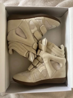 Isabel Marant beige wedge sneakers - Snygga beige sneakers från Isabel Marant med kilklack och cool design. Skorna har flera kardborreband, paneler i mocka och textil samt en brun sula. Perfekta för dig som vill ha en unik och trendig look med extra höjd. Orderbekräftelse finns och skorna är i princip nya så respektera det när ni skickar iväg prisförslag💕