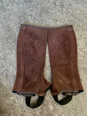 Bruna shortchaps Jacson Legacy - Bruna shortchaps i mocka och textil från Jacson Legacy. De har dragkedja och spänne upptill samt resår längs sidorna för bättre passform. Remmar under foten och detaljer i svart. Perfekta för ridning och stall.
