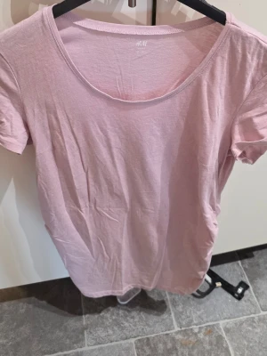 Mjuk gammelrosa mammatopp – H&M mama (Large) - Håll stilen och bekvämligheten hela vägen! Den här fina bastoppen i en mjuk, melerad gammelrosa nyans är ett oumbärligt plagg i gravidgarderoben. Med sina rynkade sidor ger den perfekt plats för magen att växa utan att toppen tappar sin snygga form. ✨  Den har en klassisk rund halsringning och korta ärmar, vilket gör den idealisk att bära som den är eller som ett mjukt lager under en kofta eller skjorta. 