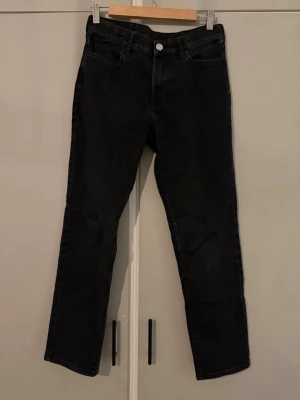 Svarta raka jeans i storlek 164 - Svarta jeans med rak passform och klassisk femficksdesign. Jeansen har knapp och dragkedja framtill samt bälteshällor i midjan. Ca 90cm.