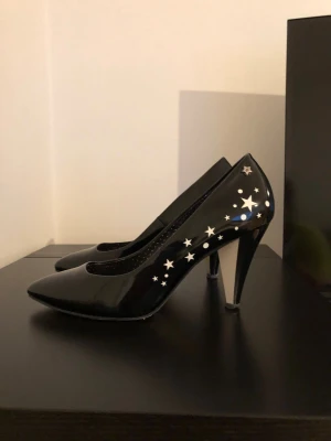 Svarta pumps med stjärnmönster Fornarina - Snygga svarta pumps från Fornarina med spetsig tå och höga klackar. Skorna har ett glansigt yttre och är dekorerade med vita stjärnor på sidan för en unik look. Perfekta för dig som vill sticka ut med coola detaljer.