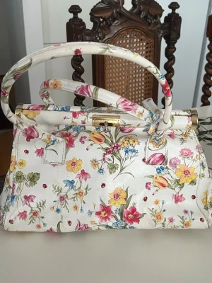  handväska med läder blommigt mönster. Genuine läder - Läder Superfin handväska  med färgglatt blommigt mönster ,. Väskan har en klassisk form, dubbla handtag och detaljer i guld.. Perfekt för dig som vill sticka ut med en unik läder accessoar.