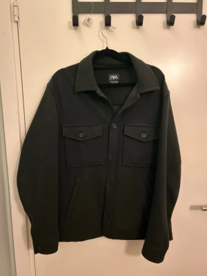 Svart overshirt från ZARA - Stilren svart overshirt från ZARA med klassisk krage, två bröstfickor med lock och knappar samt snedställda sidofickor. Overshirten har raka ärmar med knapp vid ärmslut och är tillverkad i ett mjukt, kraftigare tyg som ger en snygg siluett.