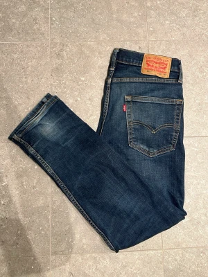 Levi's mörkblå jeans herr - Klassiska mörkblå jeans från Levi's med fem fickor, raka ben och snygga slitningar. Jeansen har den ikoniska läderpatchen bak i midjan och kontrastsömmar. Perfekta för en avslappnad och stilren look.