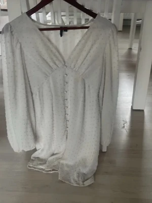 Vit långklänning med knappar - Elegant vit långklänning med v-ringad hals och dekorativa knappar framtill. Klänningen har långärmade puffärmar och ett diskret prickigt mönster. Perfekt för dig som gillar en romantisk och klassisk stil.