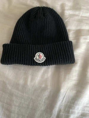 Svart ribbad mössa från Moncler - Svart ribbstickad mössa från Moncler med uppvikt kant och klassisk logotyp framtill. Perfekt accessoar för kyliga dagar och snygg till streetwear eller casual outfits.