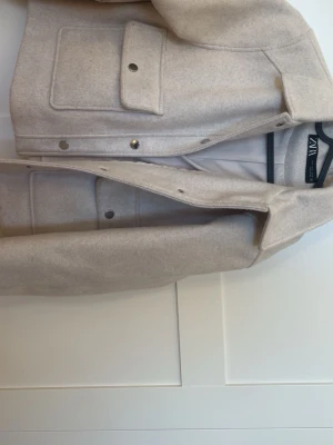 Beige overshirt från Zara - Stilren beige overshirt från Zara i mjukt ull-liknande material. Modellen har stora bröstfickor med lock och tryckknappar framtill. Perfekt lager-på-lager-plagg med klassisk krage och raka ärmar.