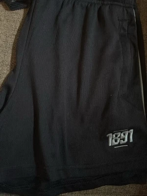 Svarta shorts med broderad 1891-logga - Svarta shorts med elastisk midja och broderad 1891-logga i vitt på ena benet. Tillverkade i ett mjukt och bekvämt material, perfekta för avslappnade dagar eller träning. Klassisk passform och diskret design.