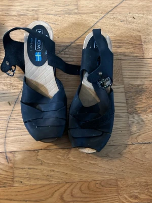 Svarta träskosandaler från Moheda - Svarta sandaler från Moheda med klassisk träbotten och breda remmar i skinn. Sandalerna har en justerbar rem med spänne runt ankeln och öppen tå. Perfekta för varma dagar och ger en stilren look med svensk design.