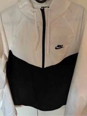 Vindjacka från NIKE - Nike vindjacka i svart och vitt med huva och dragkedja hela vägen. Jackan har två fickor med dragkedja och klassisk Nike-logga på bröstet. Tillverkad i lätt polyester, perfekt för blåsiga dagar. Snygg kontrast mellan svart och vitt, sportig och stilren look. Passar män och kvinnor därmed unisex, helt oanvänd. I storlek xl men kan passa runt 175cm.