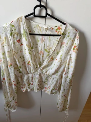 Vit blommig blus med lång ärm - Säljer en vit blus med blommigt mönster i grönt, rött och rosa. Blusen har djup v-ringning, smockad midja och långa ärmar med knytband vid ärmslut. Materialet är lätt och luftigt, perfekt för en trendig look.