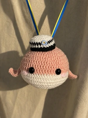 Virkad studentmaskot med mössa - Söt handvirkad maskot i rosa och vitt med svarta ögon och små armar. Maskoten har en klassisk studentmössa med blå och gula band, perfekt som accessoar eller dekoration till studenten.