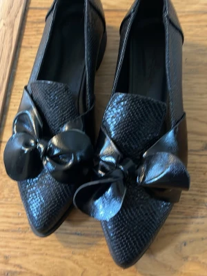 Svarta loafers med rosett och ormmönster - Snygga svarta loafers med spetsig tå, ormmönstrad yta och stora blanka rosetter framtill. Skorna har låg klack och en elegant look som sticker ut. Perfekta för dig som vill ha något extra till outfiten. 39 mer som en 38