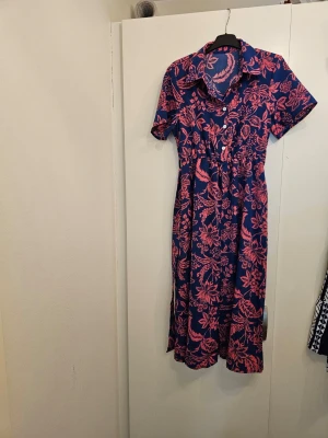Blå maxiklänning med rosa blommor - Snygg maxiklänning i blått med rosa blommönster. Klänningen har korta ärmar, knappar framtill, klassisk krage och ett matchande midjeband som markerar midjan. Materialet ser ut att vara mjukt och följsamt, perfekt för en trendig och avslappnad look.