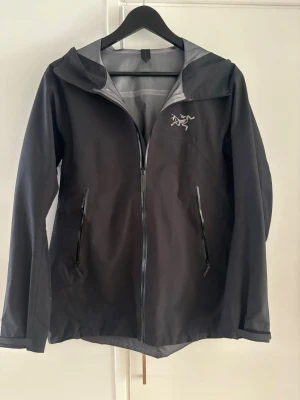 Svart regnjacka från Arc'teryx - Snygg svart regnjacka från Arc'teryx med huva och hel dragkedja framtill. Jackan har två praktiska fickor med dragkedja och diskret logga på bröstet. Tillverkad i vattentätt material, perfekt för regniga dagar.