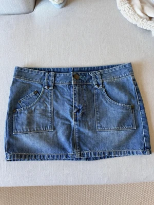 Blå jeanskjol med fickor - Snygg blå jeanskjol i klassisk modell med stora framfickor och bakfickor med dekorativ söm. Kjolen har hällor för bälte och stängs med knapp och dragkedja framtill. Perfekt till vardags för en avslappnad look.