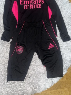 Arsenal träningsset svart/rosa Adidas - Snyggt Arsenal träningsset från Adidas i svart med rosa detaljer. Setet består av långärmad tröja med dragkedja och matchande byxor, båda med klubbmärke och Adidas-logga. Materialet är funktionsmaterial, perfekt för fotbollsträning eller gym. Endast använd fåtal gånger precis som ny säljer då den är lite för stor och gillar när de sitter tajtare den är i storlek S och jag bär vanligtvis xs!