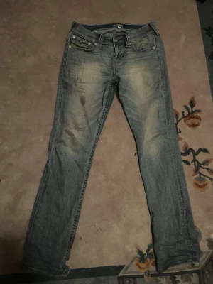 True Religion ljusblå jeans - Snygga ljusblå jeans från True Religion med klassiska kontrastsömmar och dekorativa bakfickor med lock och knappar. Jeansen har en rak passform och slitna detaljer framtill för en cool look. Perfekta för dig som gillar streetstyle.
