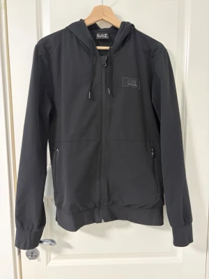 Svart windbreaker Armani - Svart hoodie från EA7 Emporio Armani med dragkedja framtill, huva med dragsko och två sidofickor med dragkedja. Minimalistisk design med logga på bröstet och ribbade muddar vid ärmslut och nederkant. Perfekt för en clean och sportig stil.