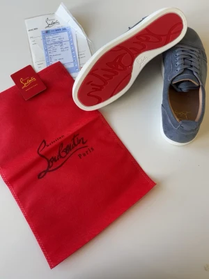 Blåa Christian Louboutin sneakers - Snygga gråa sneakers från Christian Louboutin med klassisk röd sula och vit kant. Skorna har lågt skaft, snörning och är tillverkade i mocka. Perfekta för dig som vill sticka ut med exklusiv design och ikoniska detaljer. För små för mig därför jag säljer. Ingår även box.