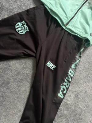 Barça träningsset från Nike, svart/turkos - Snyggt träningsset från Nike med FC Barcelona-tryck. Jackan har svart till turkos färgövergång, hel dragkedja och muddar. Byxorna är svarta med turkosa detaljer, klubbmärke och texten 'BARÇA' längs benet. Perfekt för fotbollsträning eller chill.