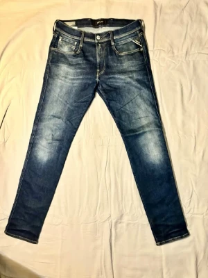 Mörkblå slim fit jeans från Replay - Säljer ett par mörkblå slim fit jeans från Replay med snygga slitningar och kontrastsömmar. Jeansen har fem fickor, dragkedja och knapp framtill samt coola detaljer på bakfickorna. Perfekta för en trendig och avslappnad look.