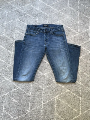 Blå slim fit jeans från Jack & Jones - Snygga blå slim fit jeans från Jack & Jones med klassisk femficksdesign och lätt slitna detaljer framtill. Jeansen har normal midja, dragkedjegylf och är tillverkade i stretchigt denim för extra komfort.