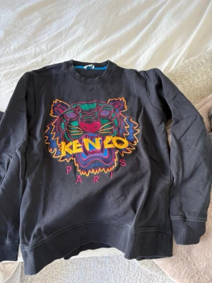 Svart Kenzo sweatshirt med tiger - Cool svart sweatshirt från Kenzo med ikoniskt broderat tigerhuvud i färgglada nyanser och texten 'KENZO PARIS' framtill. Tröjan har rund halsringning, ribbade muddar och är tillverkad i mjukt bomullsmaterial. Perfekt statement-plagg för dig som gillar streetstyle.