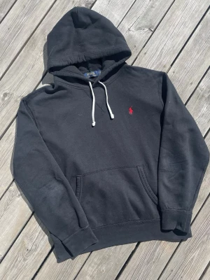 Svart hoodie från Polo Ralph Lauren - Svart hoodie från Polo Ralph Lauren med vit dragsko i huvan och klassisk röd broderad logga på bröstet. Mjuk bomullskänsla och känguruficka framtill. Perfekt för en avslappnad och stilren look.
