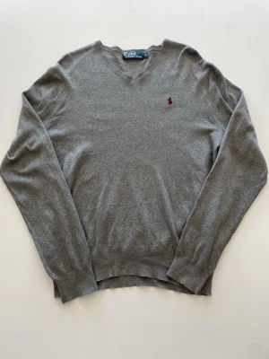 Grå V-ringad tröja Polo Ralph Lauren - Klassisk grå V-ringad tröja från Polo Ralph Lauren med den ikoniska röda loggan broderad på bröstet. Tröjan har långa ärmar och ribbade muddar vid ärmslut och nederkant. 