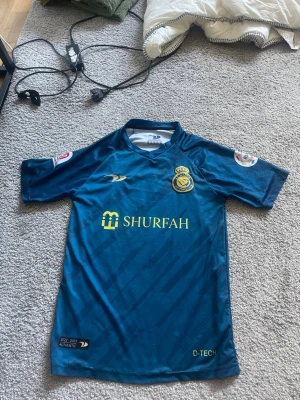 Al Nassr Ronaldo matchtröja blå - Säljer en blå Al Nassr matchtröja med Ronaldo 7 på ryggen. Tröjan har gula detaljer, klubbmärke och sponsorlogga på bröstet samt autenticitetsmärke. Korta ärmar och sportig passform, perfekt för fotbollsträning eller att bära som supporter.