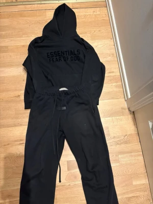 Svart hoodie Essentials Fear of God - Säljer ett svart set från Fear of God Essentials med hoodie och matchande byxor. Hoodien har stor logga på ryggen och byxorna har dragsko i midjan. Båda plaggen är i mjukt bomullsmaterial och har en avslappnad passform.