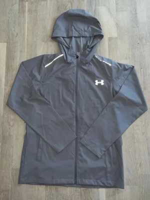 Grå vindjacka från Under Armour - Snygg grå vindjacka från Under Armour med huva och hel dragkedja. Jackan har reflekterande detaljer på axlarna och stor logga på ryggen. Perfekt för blåsiga dagar och sportiga outfits. Tillverkad i lätt och smidigt material.