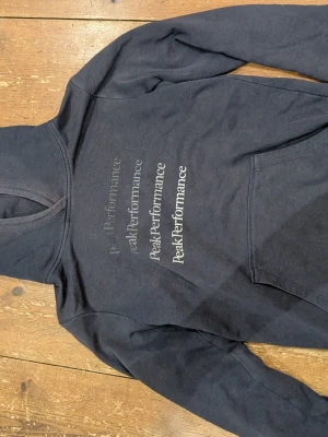 Mörkblå hoodie från Peak Performance - Snygg mörkblå hoodie från Peak Performance med tryck i vitt och grått på bröstet. Klassisk känguruficka framtill och stor huva. Perfekt för en avslappnad och sportig stil. Tillverkad i mjukt bomullsmaterial för extra komfort.