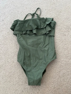 Olivgrön baddräkt med volanger - Supersnygg olivgrön baddräkt med volangdetaljer över bysten och smala axelband, kors i ryggen. Baddräkten har en enkel, klassisk passform och är tillverkad i stretchigt material som passar perfekt för strand eller pool.