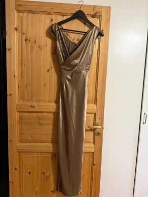 Champagnefärgad långklänning omlott - Elegant långklänning i champagnefärgat satinliknande material med omlott-skärning och v-ringad hals. Klänningen har smala axelband med snygga banddetaljer och en figurnära passform som ger en lyxig känsla. Perfekt som fest eller balklänning 