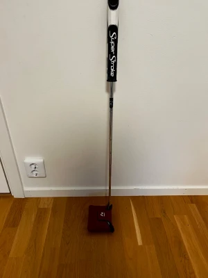 TaylorMade Spider putter SuperStroke - TaylorMade Spider putter med vitt klubbhuvud och svartvitt SuperStroke-grepp. Skaftet är i metall och ett rött headcover med Spider-logga ingår. Perfekt för golfare som vill ha stabilitet och snygg design på greenen.