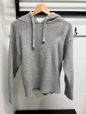 Grå stickad hoodie med huva - Mysig grå stickad hoodie med huva och dragsnören. Tröjan har ribbade muddar vid ärmslut och nederkant, samt en avslappnad passform. Perfekt för en chill och stilren look. Materialet känns mjukt och bekvämt mot huden. Lite nopprig