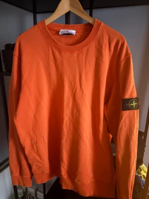 Orange sweatshirt från Stone Island - Säljer en orange sweatshirt från Stone Island med klassisk logotyp-patch på vänster ärm. Tröjan har rund halsringning och är tillverkad i mjuk bomull. Perfekt för dig som gillar streetwear och vill sticka ut med färg.