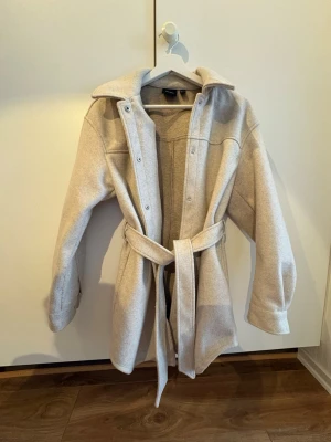 Beige kappa med bälte - Stilren beige kappa med bred krage och knytbälte i midjan. Kappan har tryckknappar framtill och är tillverkad i ett mjukt, ull-liknande material. Perfekt för kyliga dagar och ger en snygg siluett. Enkel design som passar till många outfits.