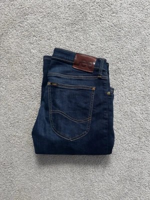 Mörkblå jeans från Lee - Snygga mörkblå jeans från Lee med klassisk femficksdesign och bruna kontrastsömmar. Jeansen har en rak passform och är tillverkade i slitstark denim med Lee-logga på läderpatchen bak i midjan. Skicket är 10/10. Storlek W31 L32. Bara att höra av dig vid frågor!!🌟