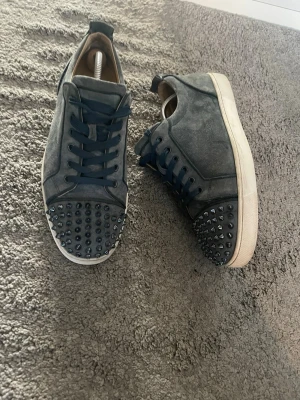 Blåa sneakers med nitar från Louboutin - Snygga blåa sneakers i mocka från Christian Louboutin med svarta nitar på tån och svarta skosnören. Skorna har en vit sula och coola detaljer som ger en edgy vibe. Perfekta för dig som vill sticka ut med din stil.